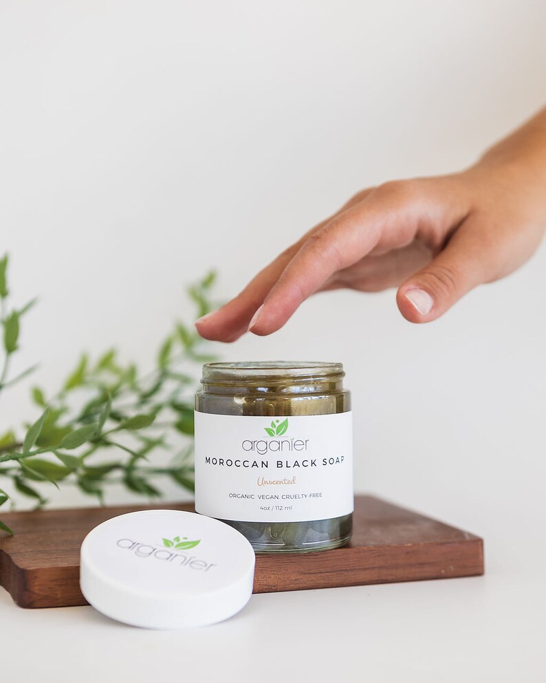 May include: A glass jar of Moroccan Black Soap with a white label. The label reads "arganier Moroccan Black Soap Unscented Organic Vegan Cruelty Free 4oz / 112 ml". The jar sits on a wooden tray with the lid beside it.