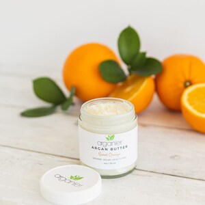 May include: A glass jar of Argan Butter with a white lid, labelled "Sweet Orange." The jar is next to whole and sliced oranges with green leaves. The product is organic, vegan, and cruelty-free.