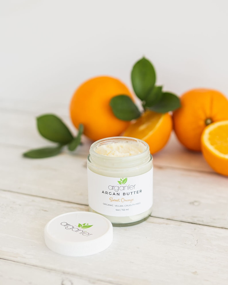 May include: A glass jar of Argan Butter with a white lid, labelled "Sweet Orange." The jar is next to whole and sliced oranges with green leaves. The product is organic, vegan, and cruelty-free.