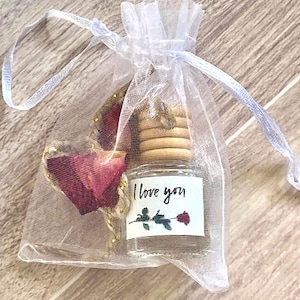 May include: A small glass bottle with a white label that says "I love you" and a small red rose illustration. The bottle is filled with a clear liquid and is surrounded by dried rose petals in a white organza bag.
