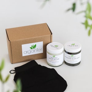 May include: A skincare set comprising a brown box with the "organier" logo, two jars of Argan Butter and Moroccan Black Soap, and a black exfoliating mitt. The jars have white lids and labels.