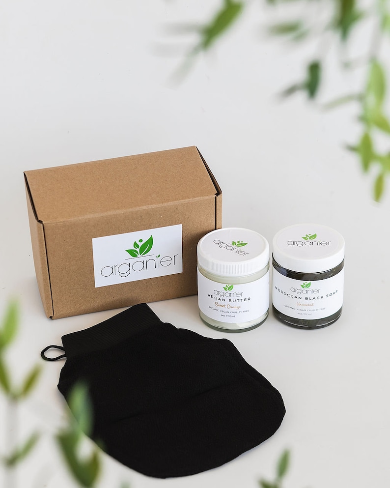 May include: A skincare set comprising a brown box with the "organier" logo, two jars of Argan Butter and Moroccan Black Soap, and a black exfoliating mitt. The jars have white lids and labels.