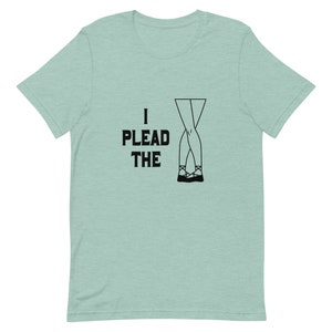 Op de afbeelding: Een lichtblauw t-shirt met de tekst "I plead the" en een zwarte lijntekening van gekruiste benen met zwarte schoenen.