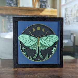 May include: A framed illustration of a green luna moth with gold accents against a dark blue background with gold crescent moons. The moth is surrounded by gold stars and green foliage.