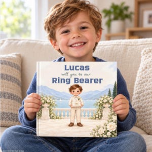 May include: A square hardback book with a white cover featuring a cartoon illustration of a young ring bearer standing on a terrace. The text on the cover reads, Lucas will you be our Ring Bearer, set against a backdrop of a lake and mountains.