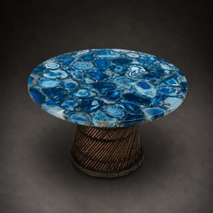 May include: A round accent table with a blue agate stone top. The tabletop is composed of irregular blue and white stone pieces. The base is a cylindrical shape with a woven, textured design in a bronze color. The table is set against a dark background.