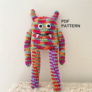 May include: A colorful crocheted monster toy with white eyes and teeth. The monster has long legs and is standing on a white fluffy surface. The text "PDF PATTERN" is visible in the upper right corner of the image.