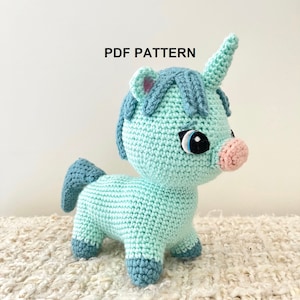 Op de afbeelding: Een gehaakt lichtblauw eenhoorn met een roze neus en blauwe ogen. Het eenhoorn staat op een wit kleed. PDF PATTERN.