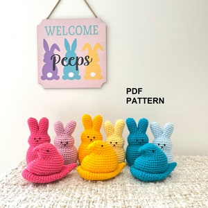 Può includere: Un set di sette peluche di coniglietto Peep all'uncinetto in vari colori pastello, tra cui rosa, viola, giallo, azzurro e turchese. I peluche sono disposti su una superficie di uncinetto bianca. Un cartello di legno con il testo "Welcome Peeps" è appeso sopra i peluche.