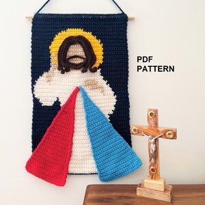 Puede incluir: Tapiz de pared tejido a crochet con una representación de Jesucristo con un halo amarillo, rodeado de una nube blanca, y un diseño triangular rojo, blanco y azul debajo. El tapiz está sujeto a una varilla de madera. El texto "PDF PATTERN" es visible en la imagen.