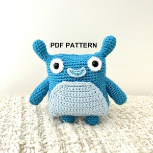 May include: A handmade blue crochet creature with a light blue belly, white eyes, and small ears. The text "PDF PATTERN" is above the creature. The toy is sitting on a white, textured surface.