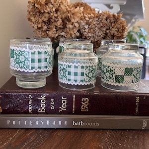 May include: A collection of clear glass jars featuring a green and white floral and gingham design. The jars are displayed on a stack of books. Each jar has a decorative scalloped edge.