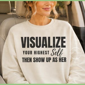 May include: A cream-colored crewneck sweatshirt featuring bold black text that reads: VISUALIZE YOUR HIGHEST Self THEN SHOW UP AS HER. The garment has long sleeves, a ribbed neckline, and a relaxed fit, providing a clean and simple aesthetic.