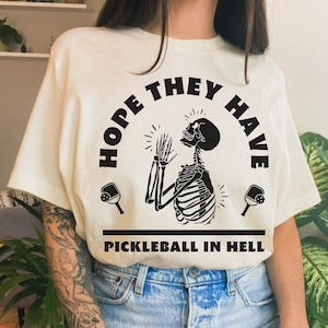 Puede incluir: Camiseta color crema con un gráfico negro de un esqueleto rezando, flanqueado por palas de pickleball. El texto dice "HOPE THEY HAVE PICKLEBALL IN HELL."
