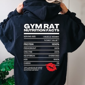 Könnte beinhalten: Ein marineblauer Hoodie mit dem Text "GYM RAT NUTRITION FACTS" in Weiß. Die Grafik listet humorvolle Ernährungsinformationen auf, darunter "Protein 1200%" und "Heartbreak Overdosed". Eine rote Lippen-Grafik ist ebenfalls vorhanden.
