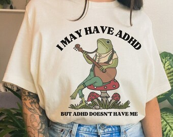 Può includere: Maglietta bianca con una rana verde che suona un banjo e seduta su un fungo. Il testo sulla maglietta recita "I may have ADHD but ADHD doesn't have me."