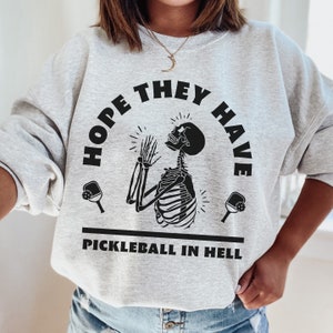 Puede incluir: Sudadera gris claro con el texto negro "HOPE THEY HAVE PICKLEBALL IN HELL". El diseño presenta un esqueleto con las manos levantadas y palas de pickleball a cada lado.