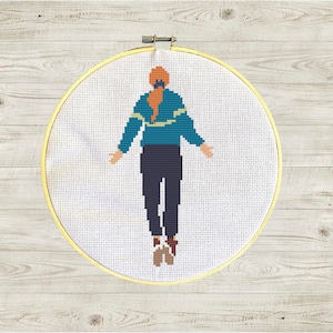 May include: Cross-stitch embroidery hoop with a colorful illustration of a person wearing a teal sweater and blue pants.
