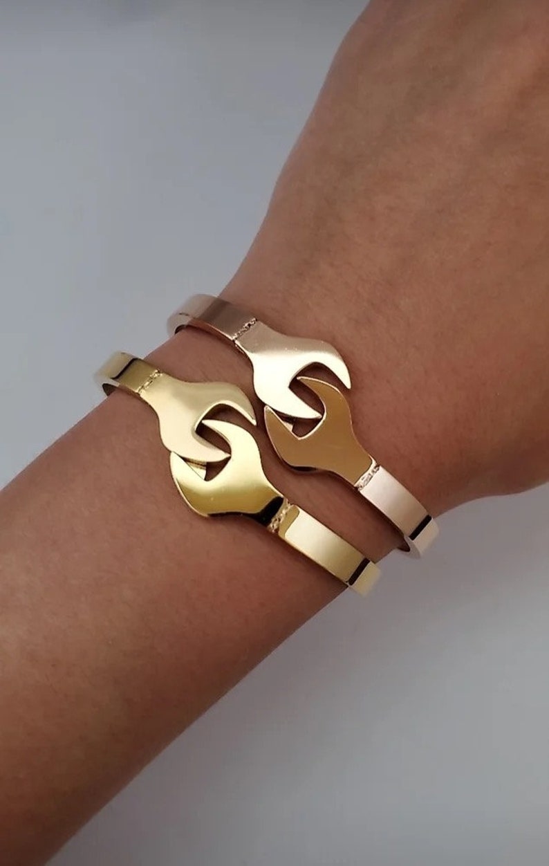 Puede incluir: Dos pulseras doradas con un dise&ntilde;o de llave inglesa. Las pulseras est&aacute;n apiladas una encima de la otra en la mu&ntilde;eca de una persona.