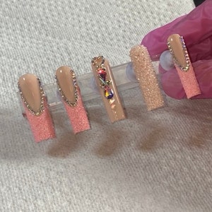 May include: Five long, nude-coloured artificial nails with pink glitter tips. The nails are arranged on a white surface and are decorated with rhinestones and a gold chain.