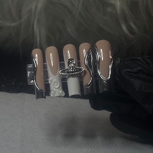 May include: Five nude-colored fake nails with white tips and silver accents. The nails are arranged on a black display card. The middle nail has a small, silver orb with a white orb in the center.