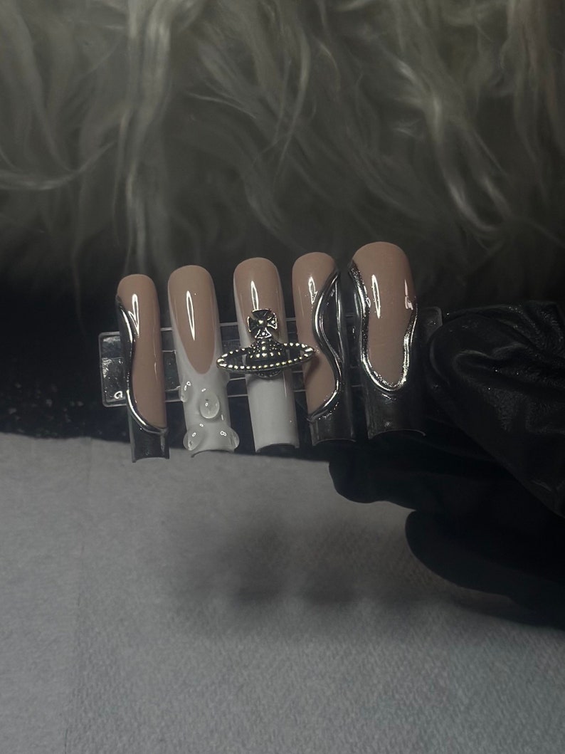 May include: Five nude-colored fake nails with white tips and silver accents. The nails are arranged on a black display card. The middle nail has a small, silver orb with a white orb in the center.