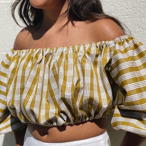 May include: Mustard yellow and white plaid off-the-shoulder crop top with short sleeves. The top is paired with white linen pants. The fabric has a textured appearance. The model is standing in front of a white wall.