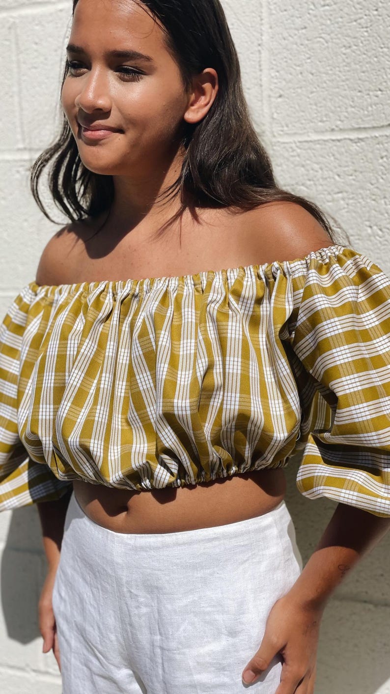 May include: Mustard yellow and white plaid off-the-shoulder crop top with short sleeves. The top is paired with white linen pants. The fabric has a textured appearance. The model is standing in front of a white wall.