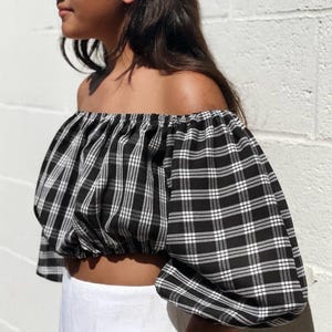 May include: Off-the-shoulder crop top in a black and white plaid pattern. The top features puffed sleeves and a gathered neckline. The fabric has a classic checkered design. The top is paired with white bottoms.