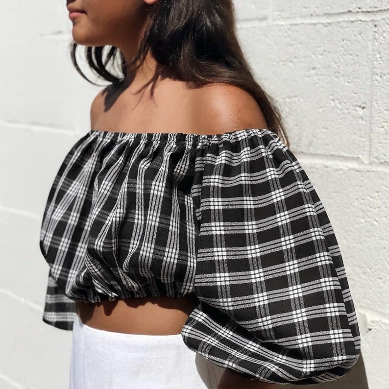 May include: Off-the-shoulder crop top in a black and white plaid pattern. The top features puffed sleeves and a gathered neckline. The fabric has a classic checkered design. The top is paired with white bottoms.