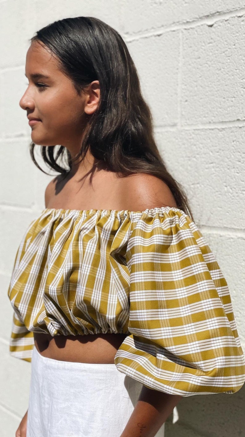 May include: Mustard yellow and white plaid off-the-shoulder crop top with puffy sleeves. The top is made of a lightweight fabric and has an elastic neckline. The model is wearing a white skirt. Fashion apparel.
