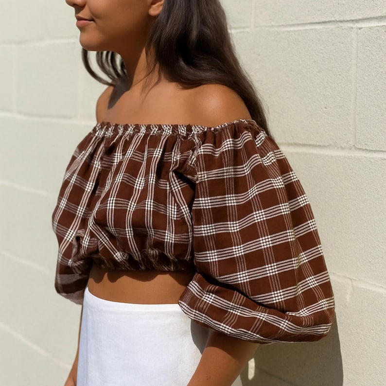 May include: An off-the-shoulder crop top in a brown and white plaid pattern. The top has puffy sleeves and an elastic neckline. The bottom half of the image shows a white skirt. The top is made of a lightweight fabric.
