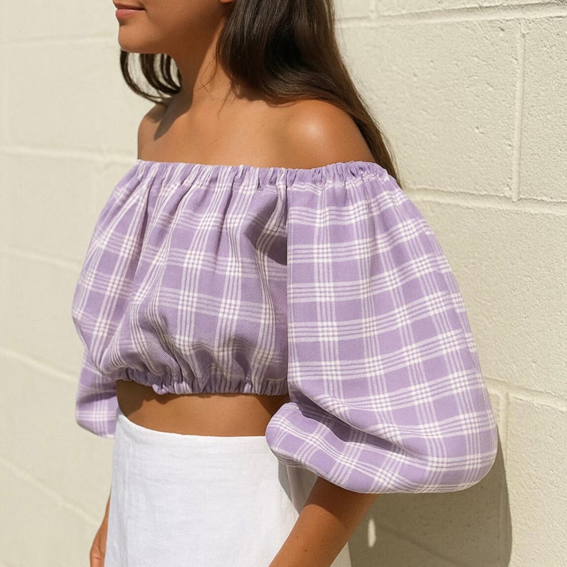 May include: A lilac off-the-shoulder crop top with a plaid pattern. The top features puffed sleeves and a gathered neckline. The fabric is a light purple with a white grid pattern. The top is paired with a white skirt.