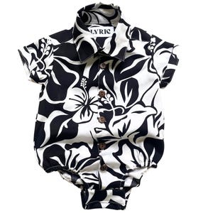 May include: Black and white baby onesie with a floral print and brown buttons. The onesie has a collar and short sleeves. The text "LYRIC" is printed on the collar.