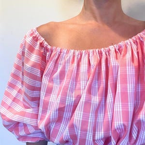 May include: A pink and white plaid off-the-shoulder top. The top has a gathered neckline and puffy sleeves. The fabric is a light cotton, and the pattern is a classic gingham check. The top is a stylish and comfortable garment.