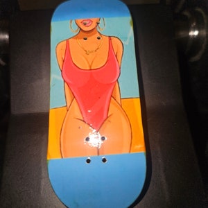 May include: A blue and orange fingerboard with a graphic of a person wearing a red swimsuit. The fingerboard has two black wheels.