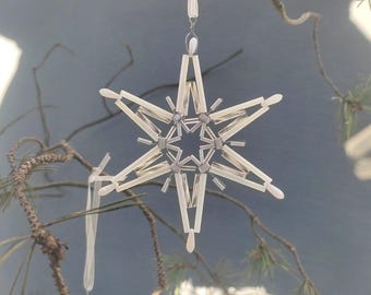 May include: A white beaded star ornament with a silver wire loop for hanging. The star is made of small, white beads and has a delicate, intricate design.