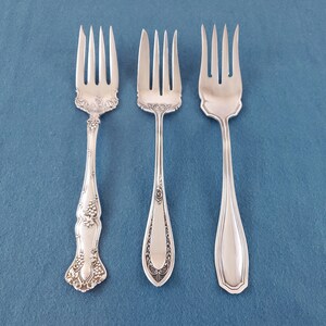May include: Three silver-toned metal forks arranged side by side. The left fork features an ornate grape and vine relief pattern. The center fork has an intricate scrollwork design on the handle. The right fork has a smooth, polished finish.