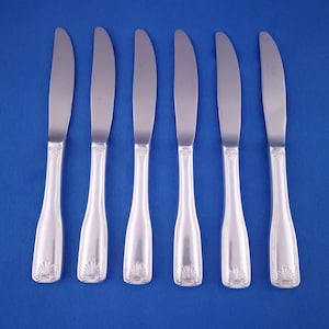 May include: Six silver butter knives arranged in a row on a blue background. Each knife has a curved blade and a decorative handle with a shell-like design at the base. The knives are made of a shiny, reflective metal.