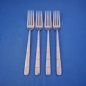 May include: Four stainless steel forks with a geometric design on the handles, arranged against a solid blue background. The forks have tines and a rectangular pattern on the handle.