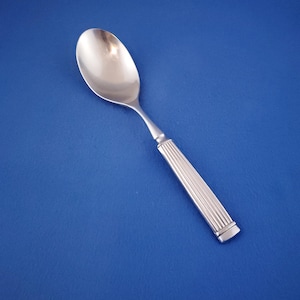 May include: A silver spoon with a rounded bowl and a handle featuring vertical grooves. The spoon is set against a solid blue background, highlighting its polished surface and classic design. Ideal for serving or eating.