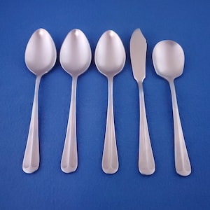 May include: A set of five stainless steel silverware pieces, including three spoons, a butter knife, and a serving spoon, arranged on a blue surface. The spoons have oval bowls and elongated handles. The butter knife has a rounded blade.