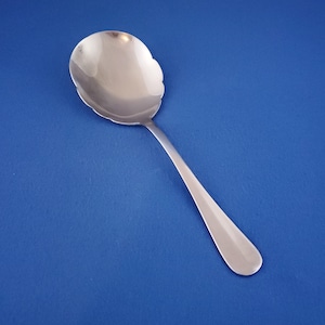 May include: A silver serving spoon with a scalloped edge bowl and a long, slender handle. The spoon is made of polished metal and is set against a solid blue background.