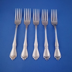 May include: Five silver forks with ornate handles and tines are arranged on a blue background. The handles have a floral design at the base. The forks are evenly spaced and angled slightly.
