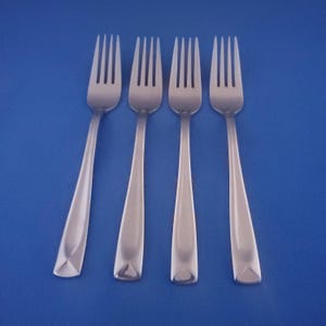 May include: Four stainless steel forks arranged on a blue background. Each fork has a long, tapered handle and four tines. The handles have a geometric design at the base. The forks are evenly spaced and angled slightly.