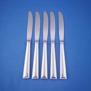 May include: Five silver butter knives arranged vertically against a solid blue background. Each knife has a rounded blade and a handle that tapers from a rectangular base to a narrower top. The knives are evenly spaced and reflect the light.