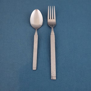 May include: A stainless steel spoon and fork set. The spoon has an oval bowl, and the fork has four tines. Both utensils have textured handles. The set is displayed on a blue background.