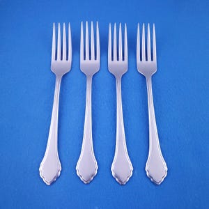 May include: Four stainless steel forks with a decorative handle design, arranged on a blue background. Each fork has four tines and a scalloped edge at the base of the handle. The forks are evenly spaced and reflect light.