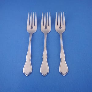 May include: Three stainless steel forks with a decorative handle and a diamond-shaped cutout in the tines. The forks are arranged on a solid blue background, showcasing their elegant design. Ideal for formal dining settings.