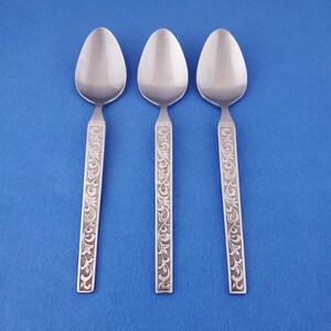 May include: Three stainless steel spoons with a floral engraved design on the handles. The spoons have an oval bowl shape and are set against a solid blue background. The spoons are a part of a flatware set.